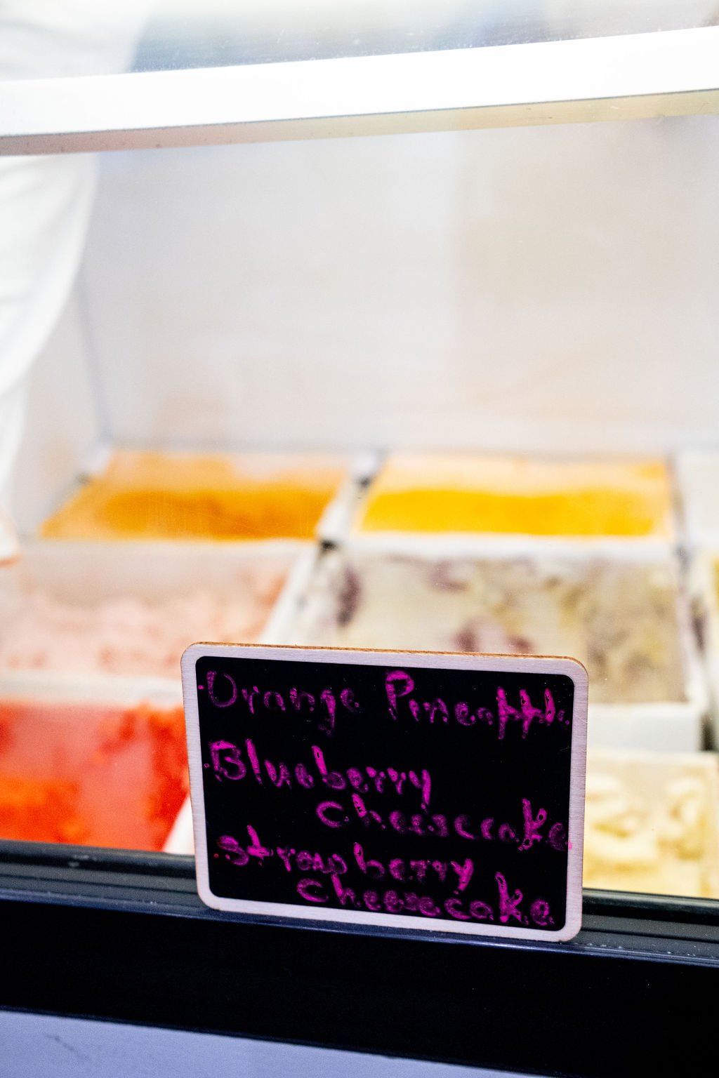 An ice cream display case with a handwritten chalkboard sign listing flavors like Orange Pineapple and Cheesecake.