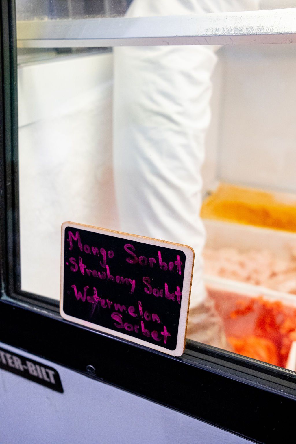 A small chalkboard sign on a freezer case lists three sorbet flavors: Mango, Strawberry, and Watermelon.
