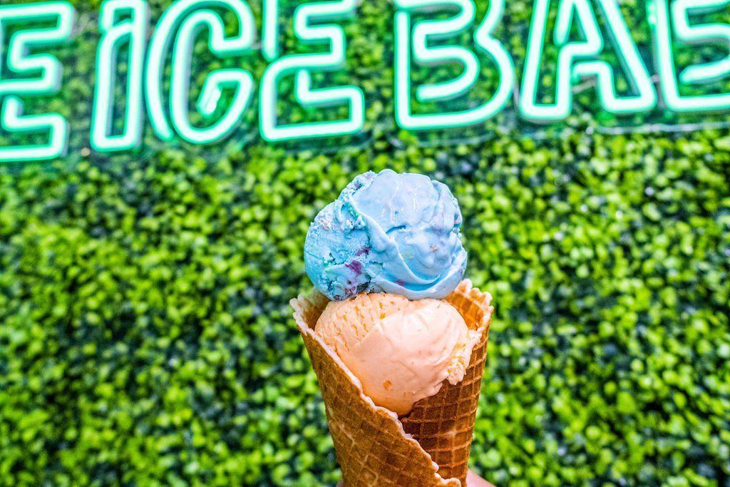 A two-scoop ice cream cone, one blue and one orange, held in front of a green wall with neon signage.