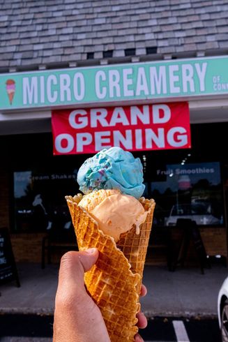 A hand holds a double-scoop ice cream cone with blue and orange scoops in front of a Micro Creamery shop.