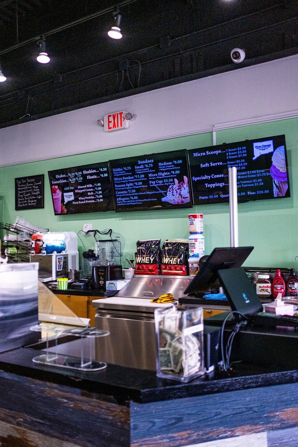 A shop counter with black equipment, a tip jar, and digital menu boards mounted on a light green wall under an exit sign.
