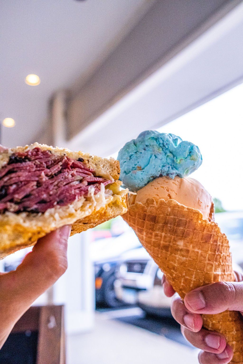 Two hands hold a pastrami sandwich and a blue-topped waffle cone ice cream against a blurred outdoor setting.