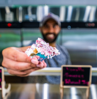 Co-owner of ice cream shop holding a small plastic spoon with a scoop of purple and white ice cream toward the camera.