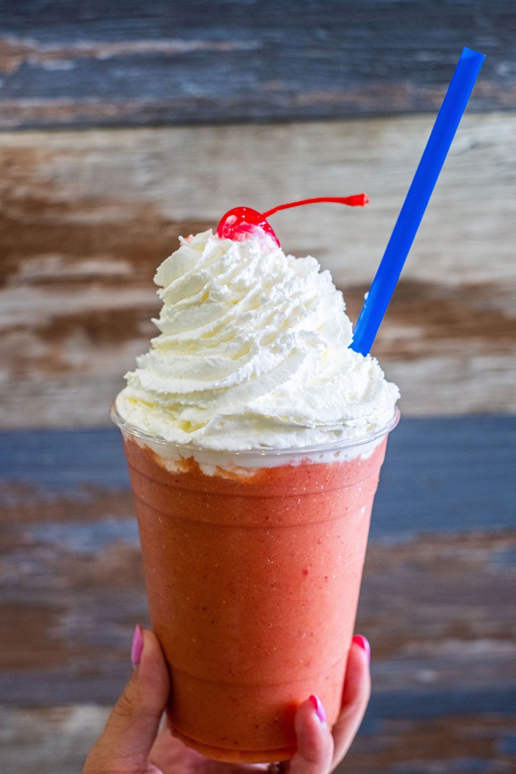 A hand holds a pink smoothie in a plastic cup, topped with whipped cream, a cherry, and a blue straw against wood siding.