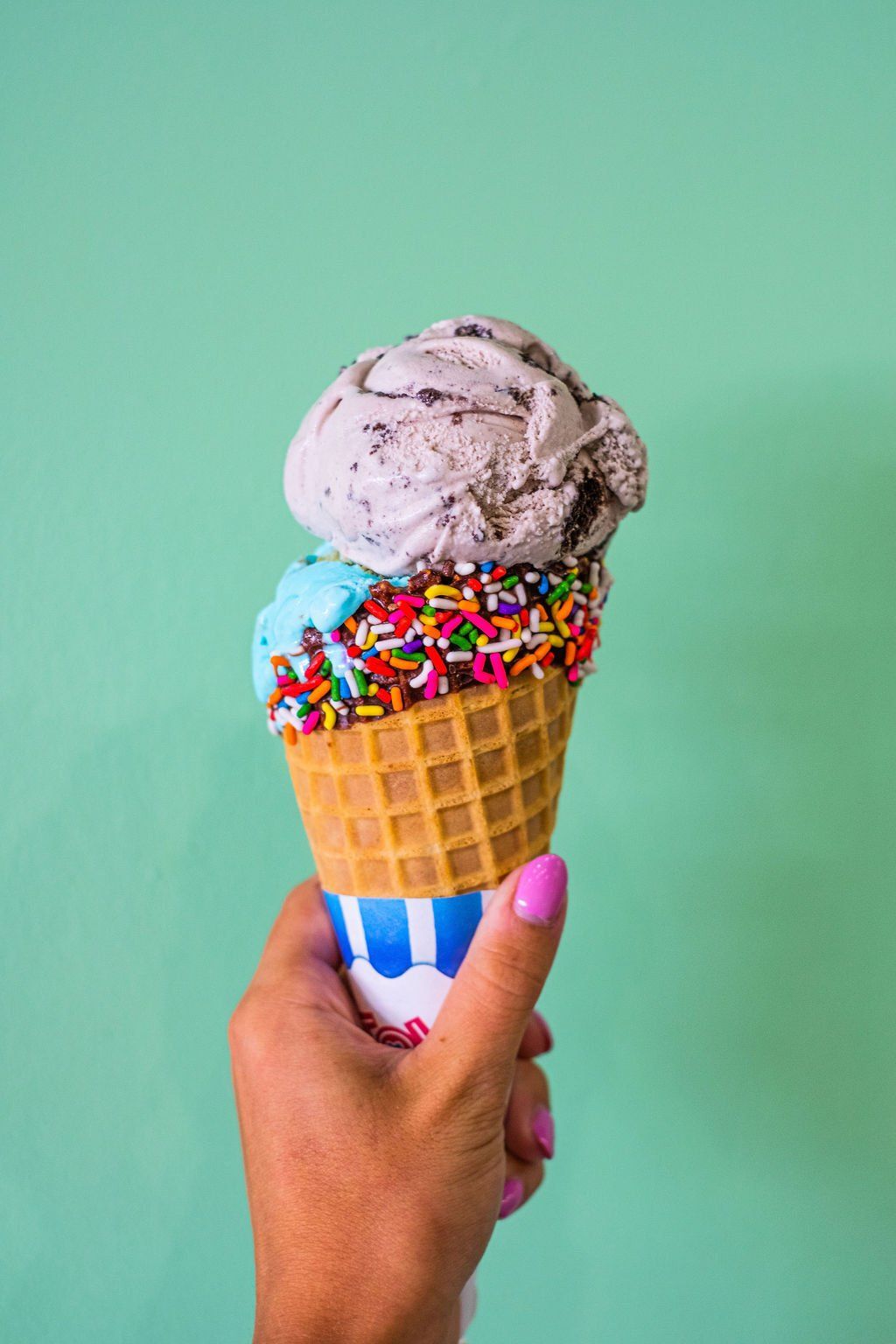 A hand holds a waffle cone topped with a scoop of cookies and cream ice cream and colorful rainbow sprinkles.