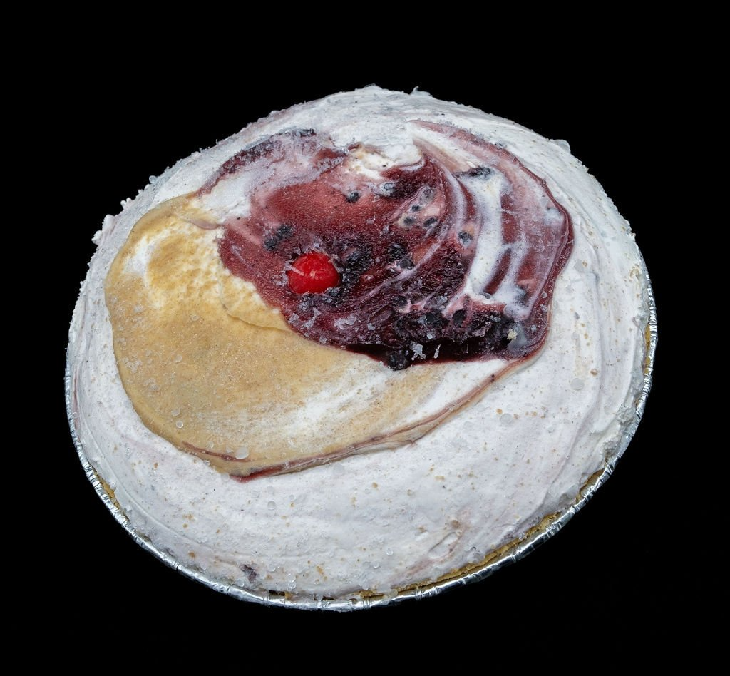 A round dessert in a foil pan, topped with white cream, a dark berry swirl, and a single bright red cherry.