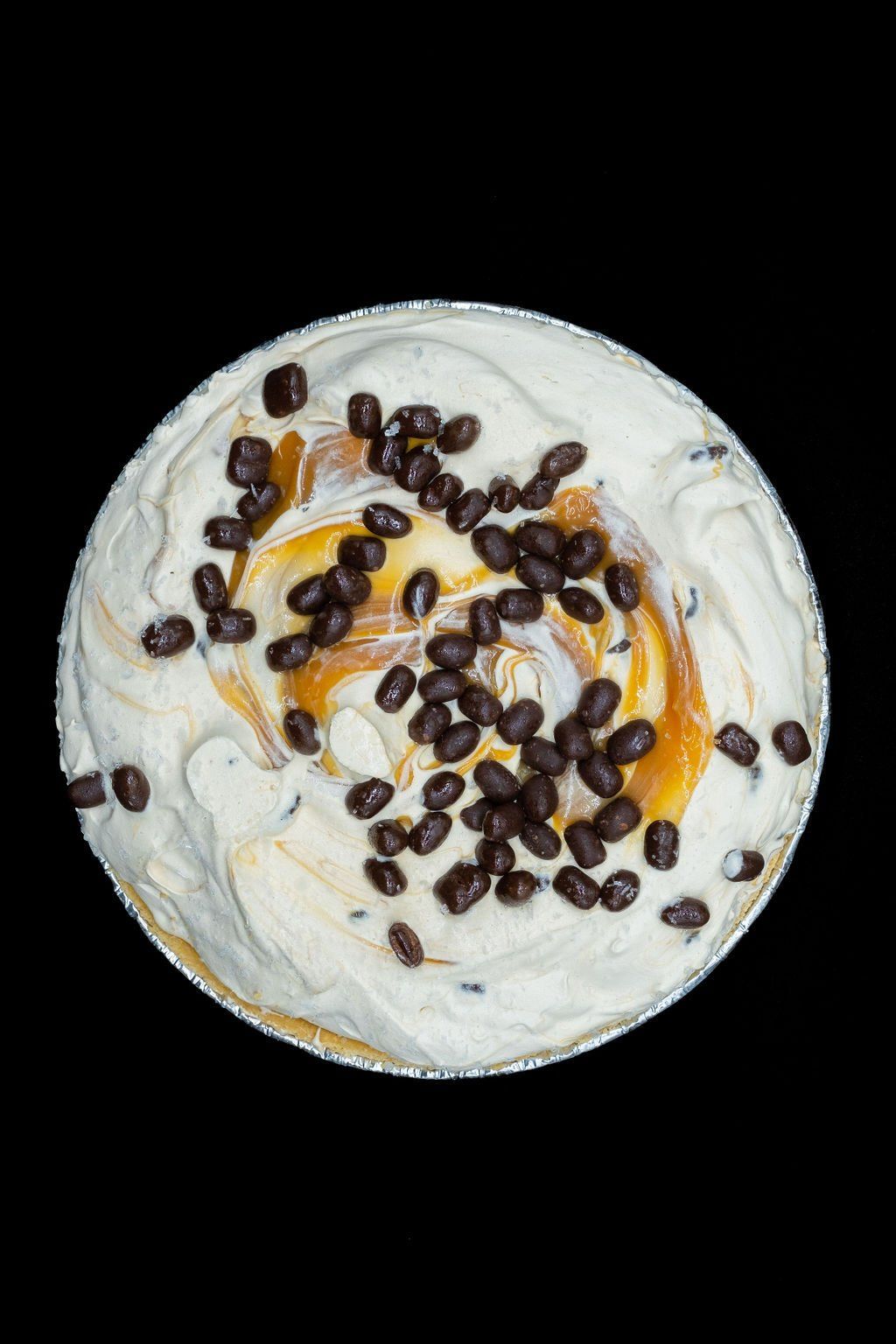 A round pie with creamy white topping, caramel swirls, and dark chocolate-covered espresso beans on a black background.