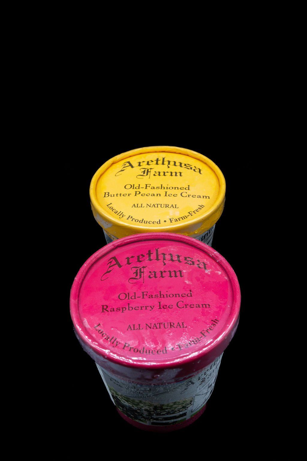 Two Arethusa Farm ice cream pints stacked against a black background: a red Raspberry and a yellow Butter Pecan.