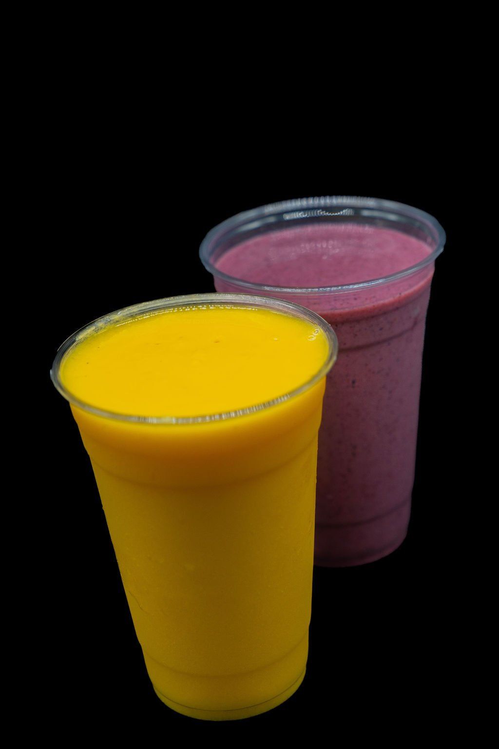 Two clear plastic cups of blended fruit smoothies, one bright yellow and one deep purple, set against a black background.