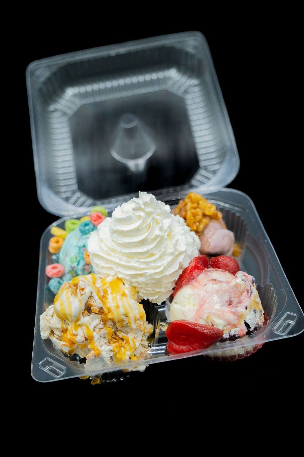 A plastic container holds four scoops of ice cream topped with whipped cream, colorful cereal, and sliced strawberries.