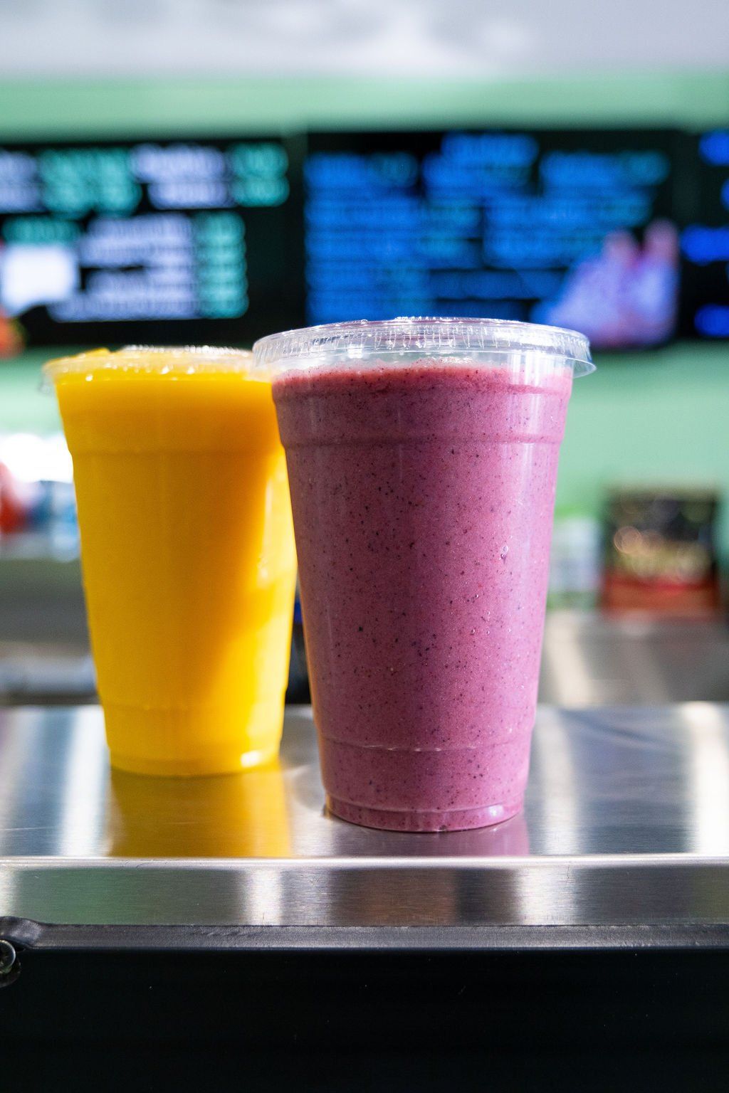 Two plastic cups with fruit smoothies, one yellow and one purple, sit on a reflective metal counter in front of a menu.