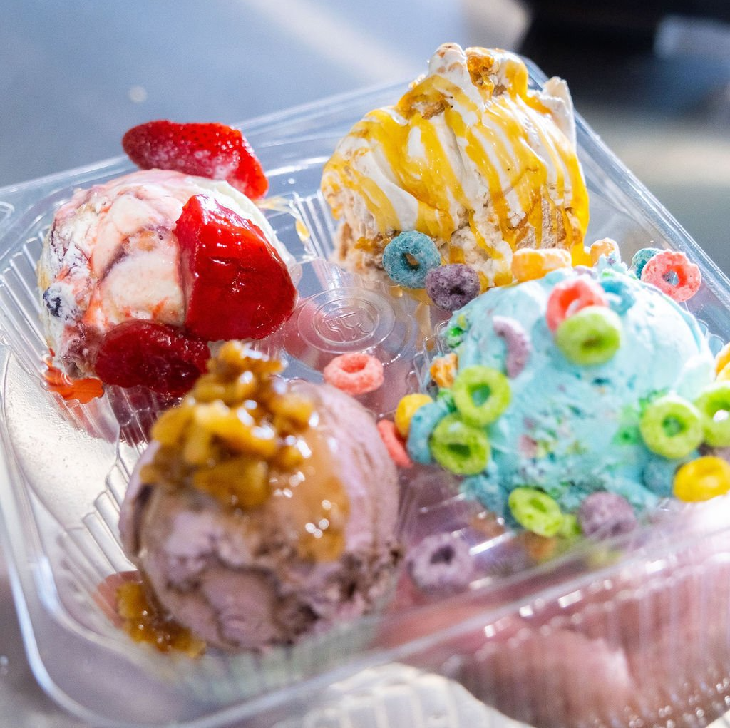 A plastic container holds four scoops of ice cream with strawberry, caramel, walnut, and colorful cereal toppings.