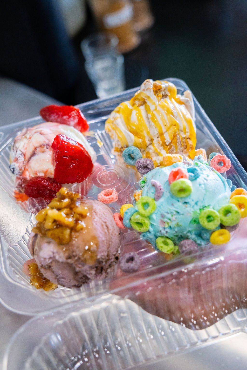 A clear plastic container holding four scoops of colorful ice cream topped with strawberries, caramel, and cereal loops.