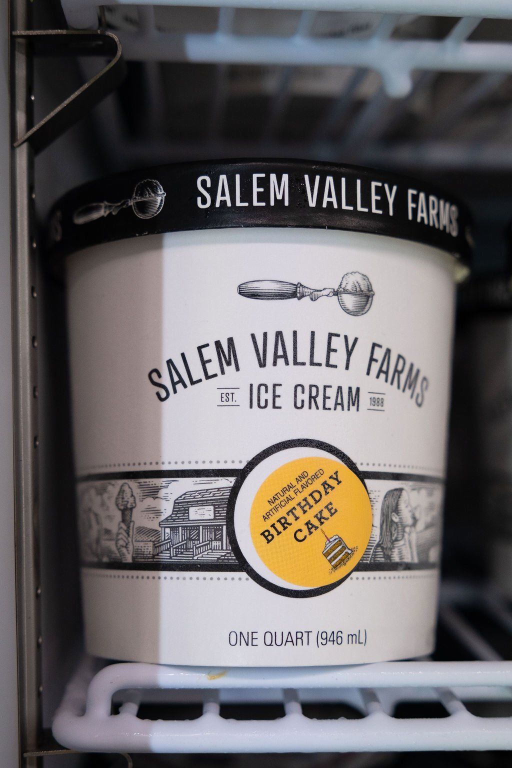 A quart container of Salem Valley Farms Birthday Cake ice cream sitting on a wire shelf inside a freezer.