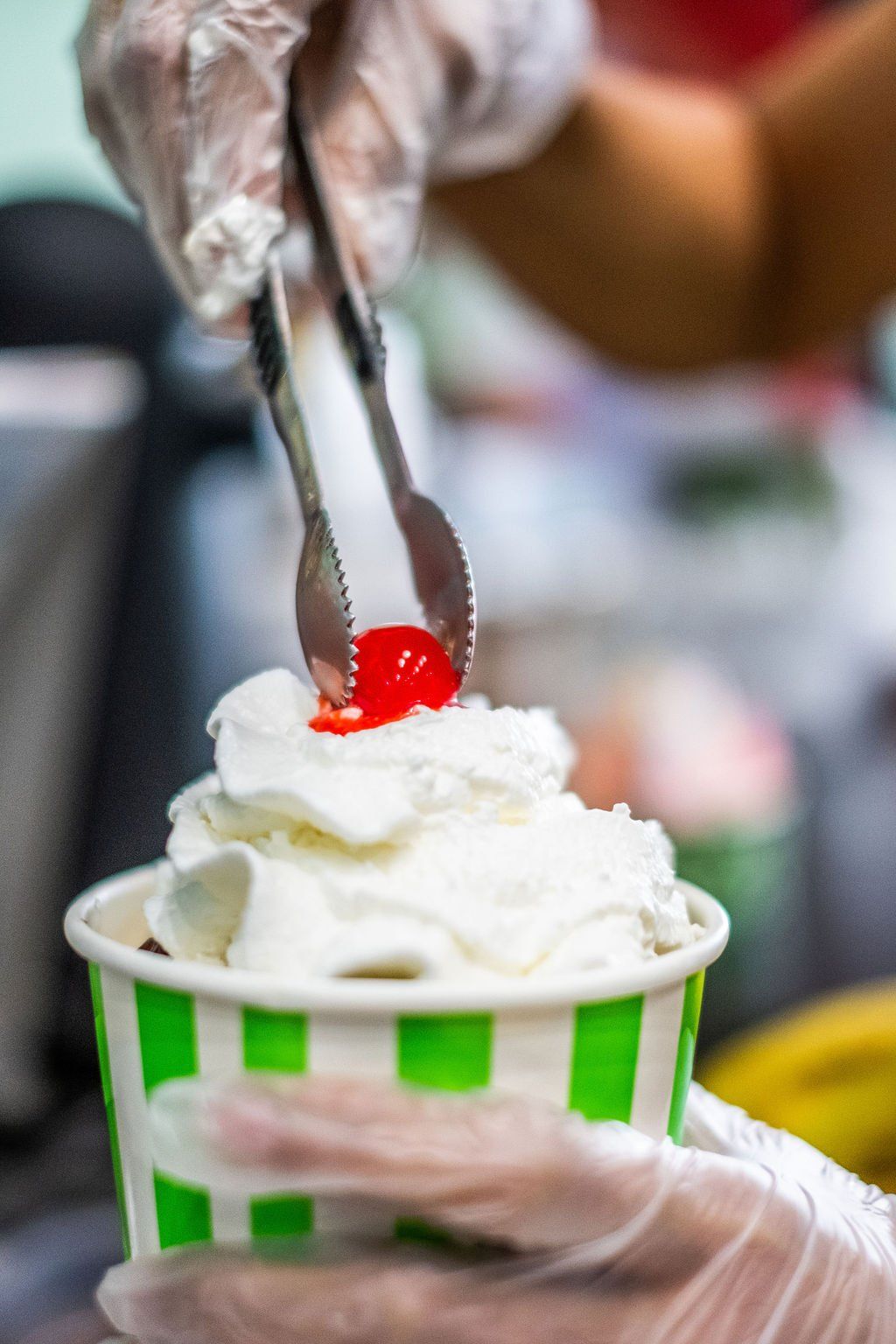 A gloved hand uses tongs to place a bright red cherry atop a mound of whipped cream in a green-striped paper cup.