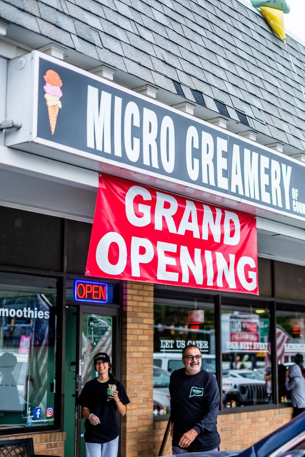 A person walks from a Micro Creamery shop during a Grand Opening event, with a red banner hanging from the storefront.