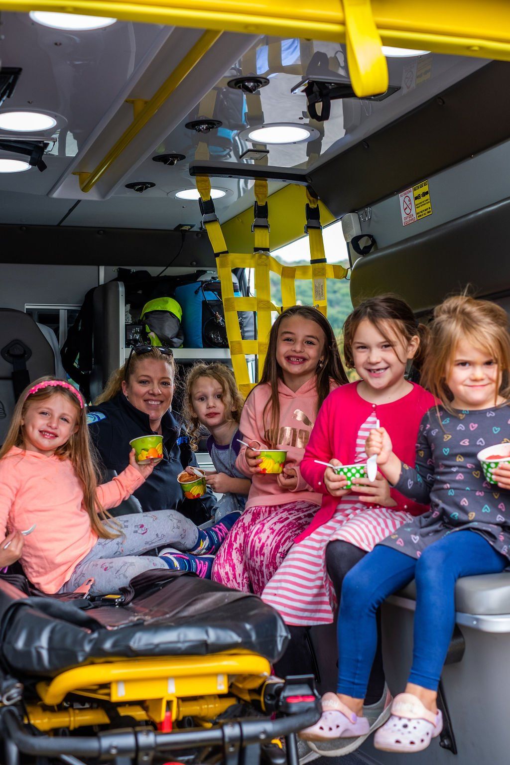 Children in a vehicle, sitting near a gurney and holding small cups, smiling at the camera.