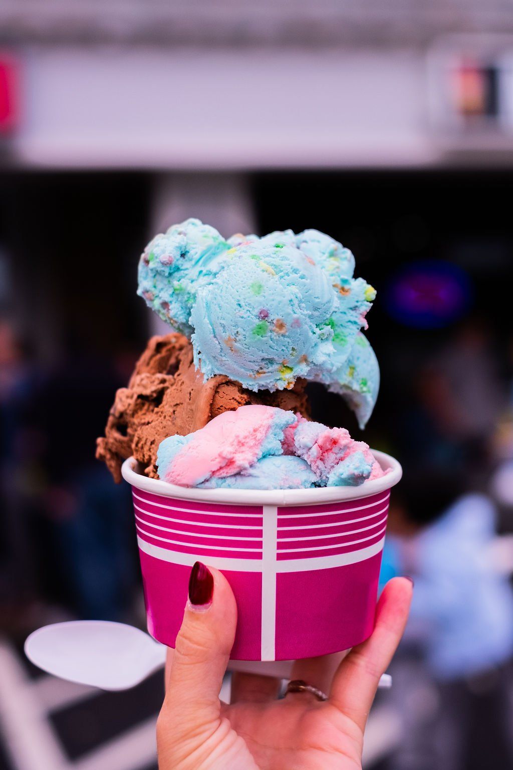 A hand holds a magenta paper cup filled with three scoops of colorful ice cream against a blurred outdoor background.