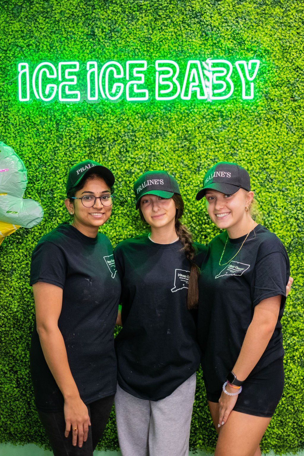 Three smiling people in matching black shirts and hats stand before a green wall with the neon sign 