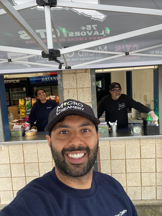 A smiling man in a baseball cap takes a selfie in front of a concession stand with two staff members working inside.
