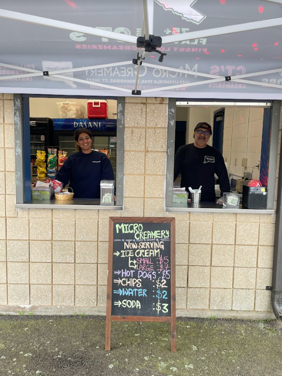 A person stands at each of two windows serving an ice cream and food stand, with a menu board displayed on the ground.