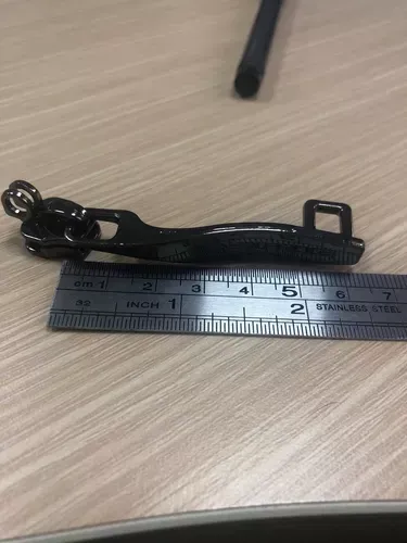 Tirador de cremallera de metal negro junto a una regla sobre una mesa de madera clara.