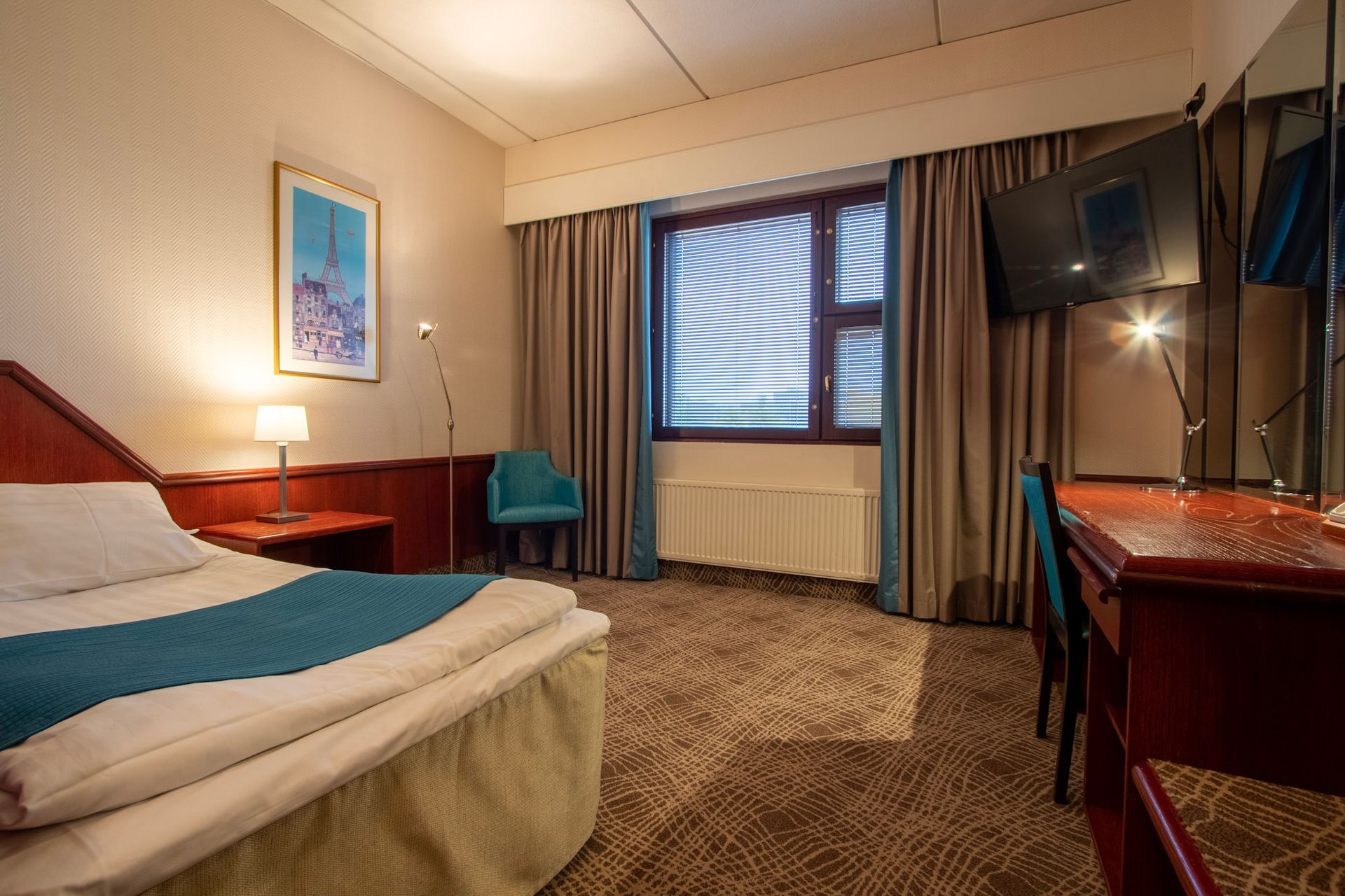 A hotel room with a single bed, wooden desk and chair, blue accent chair, and large window with tan curtains.