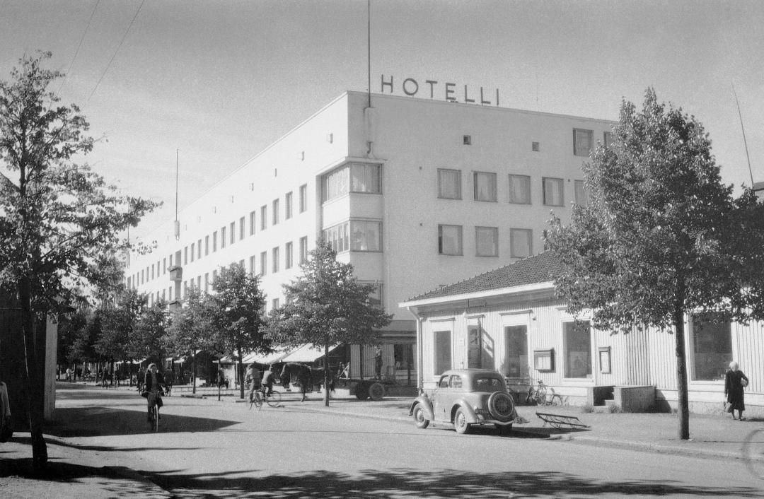 Mustavalkoinen katunäkymä modernista hotellirakennuksesta vanhemman, pienemmän rakennuksen ja pysäköidyn auton vieressä.
