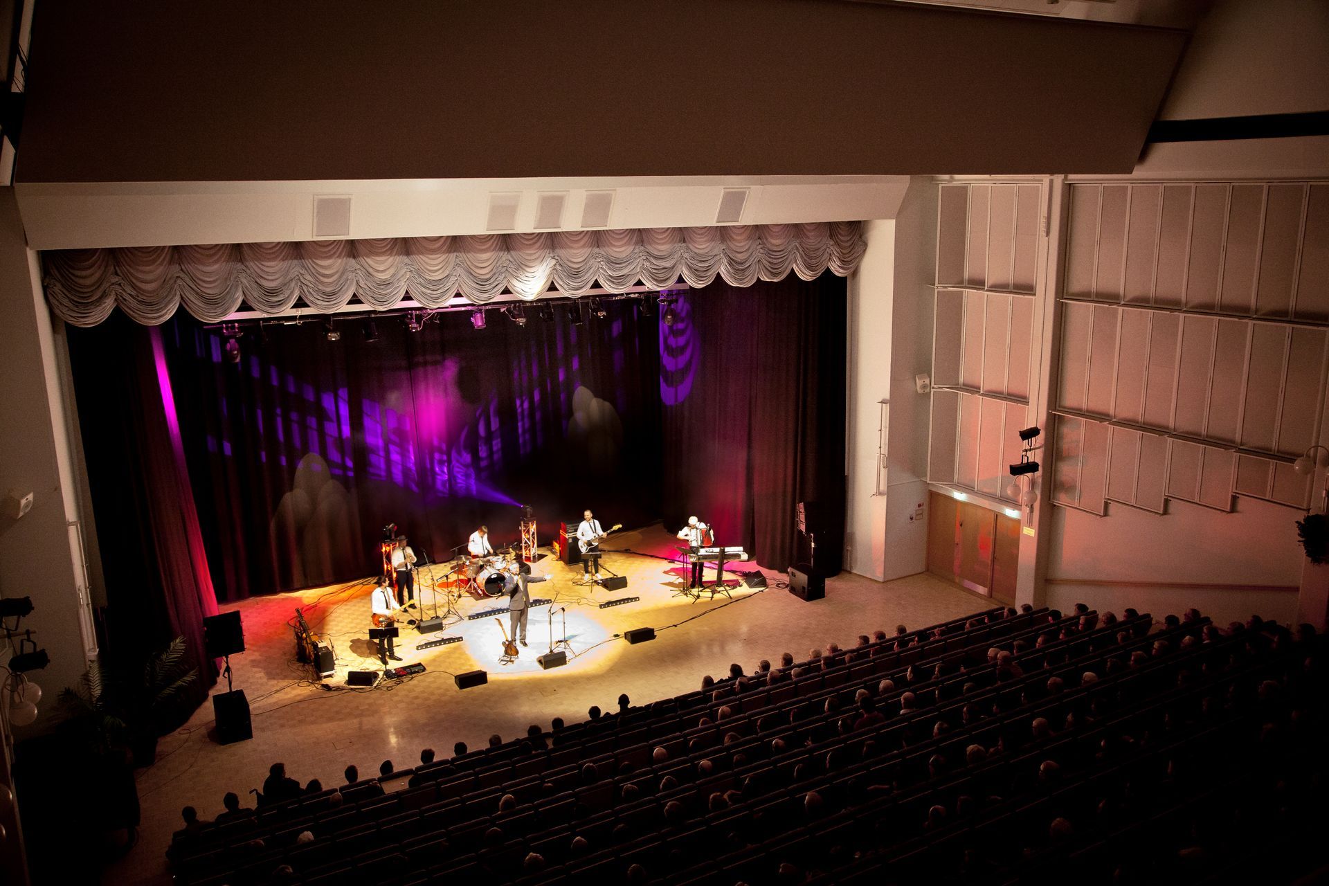 Ylhäältä kuvattu kuva bändistä esiintymässä valaistussa teatterilavalla istuvan yleisön edessä hämärästi valaistussa auditoriossa.