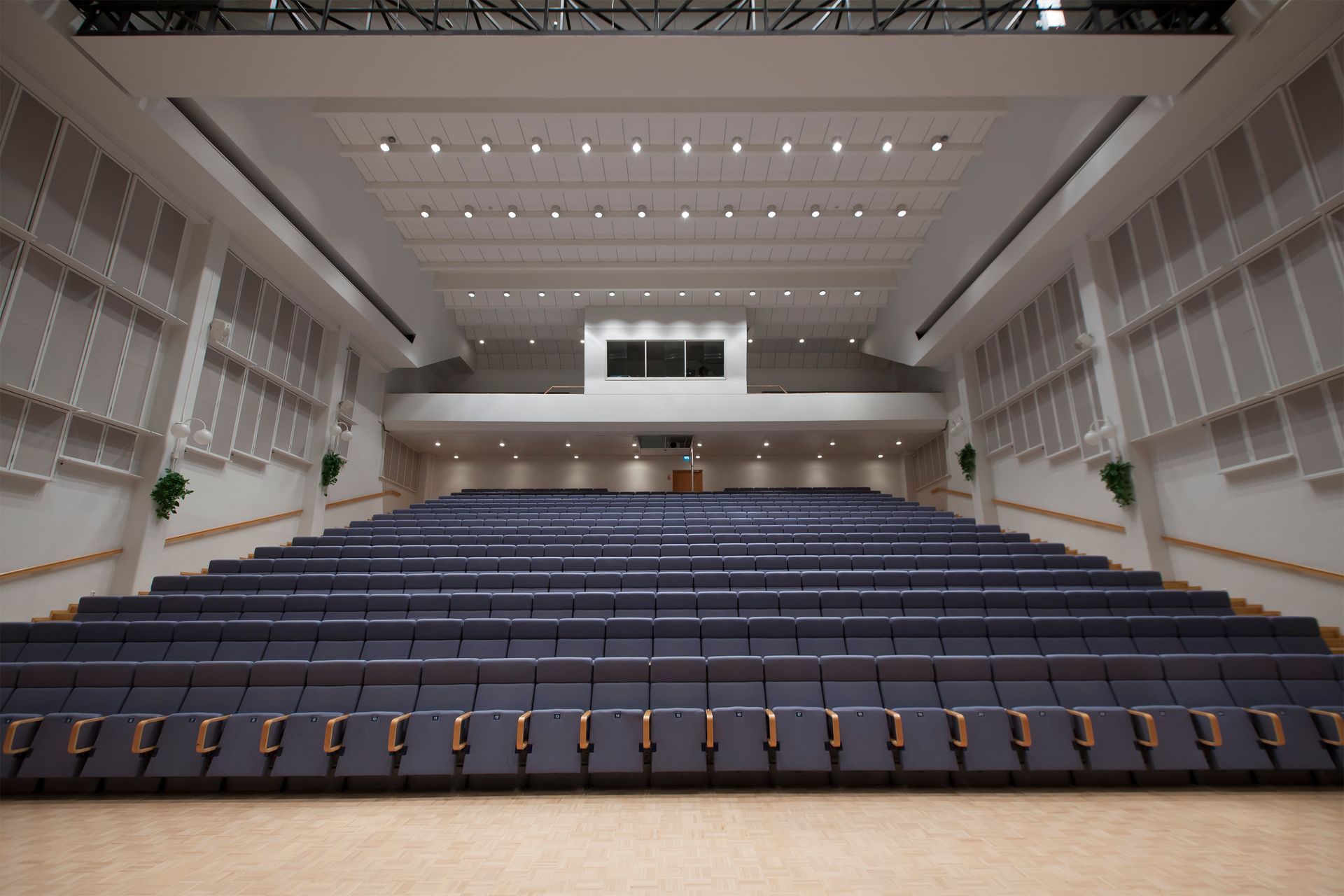 Laajakuvanäkymä suuresta, tyhjästä auditoriosta, jossa on rivejä sinisiä porrastettuja istuimia vastapäätä kirkkaissa valoissa valaistua puista lavaa.