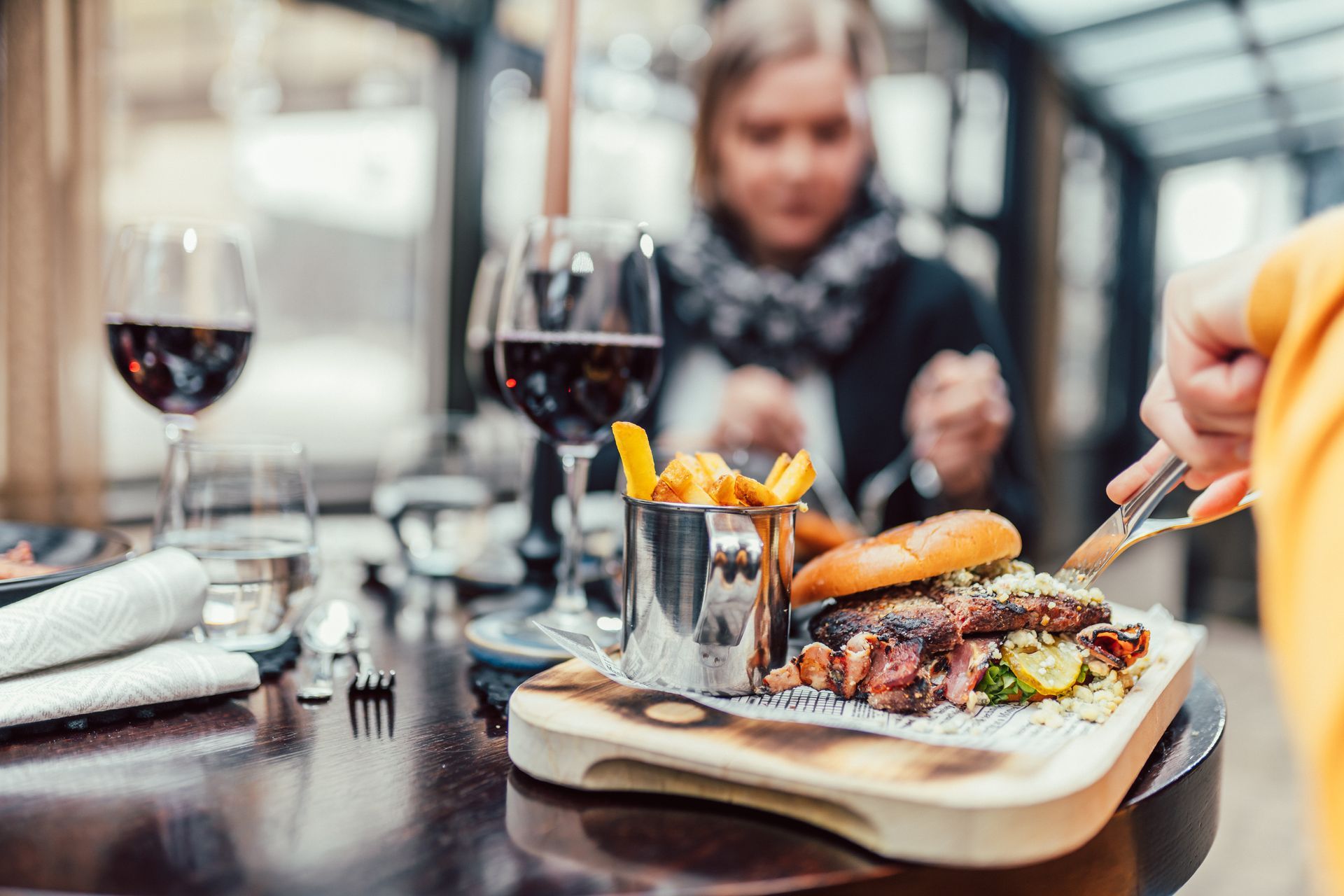 A person uses a knife and fork to eat a burger and fries from a wooden board at a table with glasses of red wine.