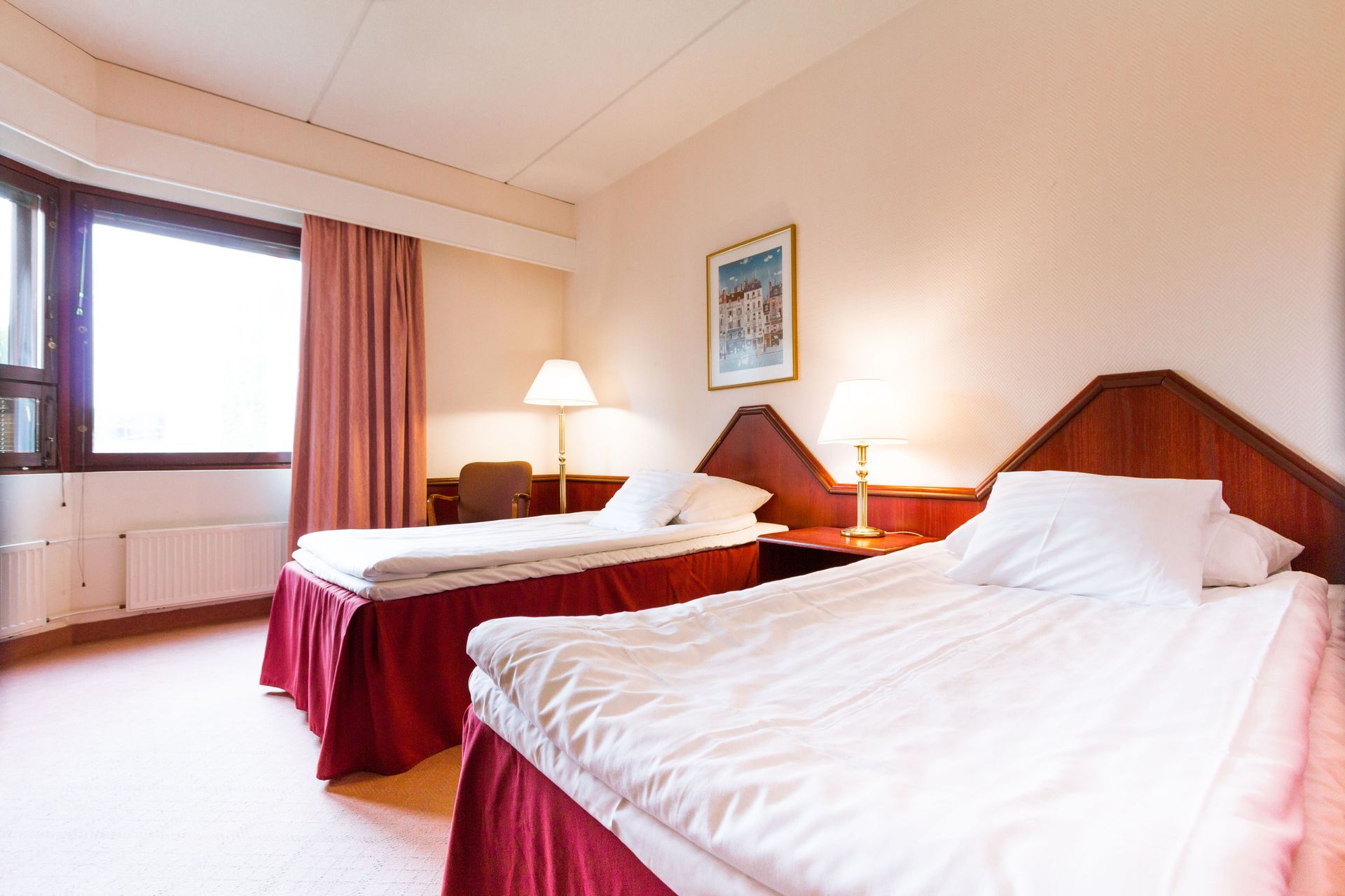 Hotellihuone, jossa on kaksi erillistä sänkyä, valkoiset liinavaatteet, punaiset hameet, puiset sängynpäädyt ja yölamput maalauksen alla.
