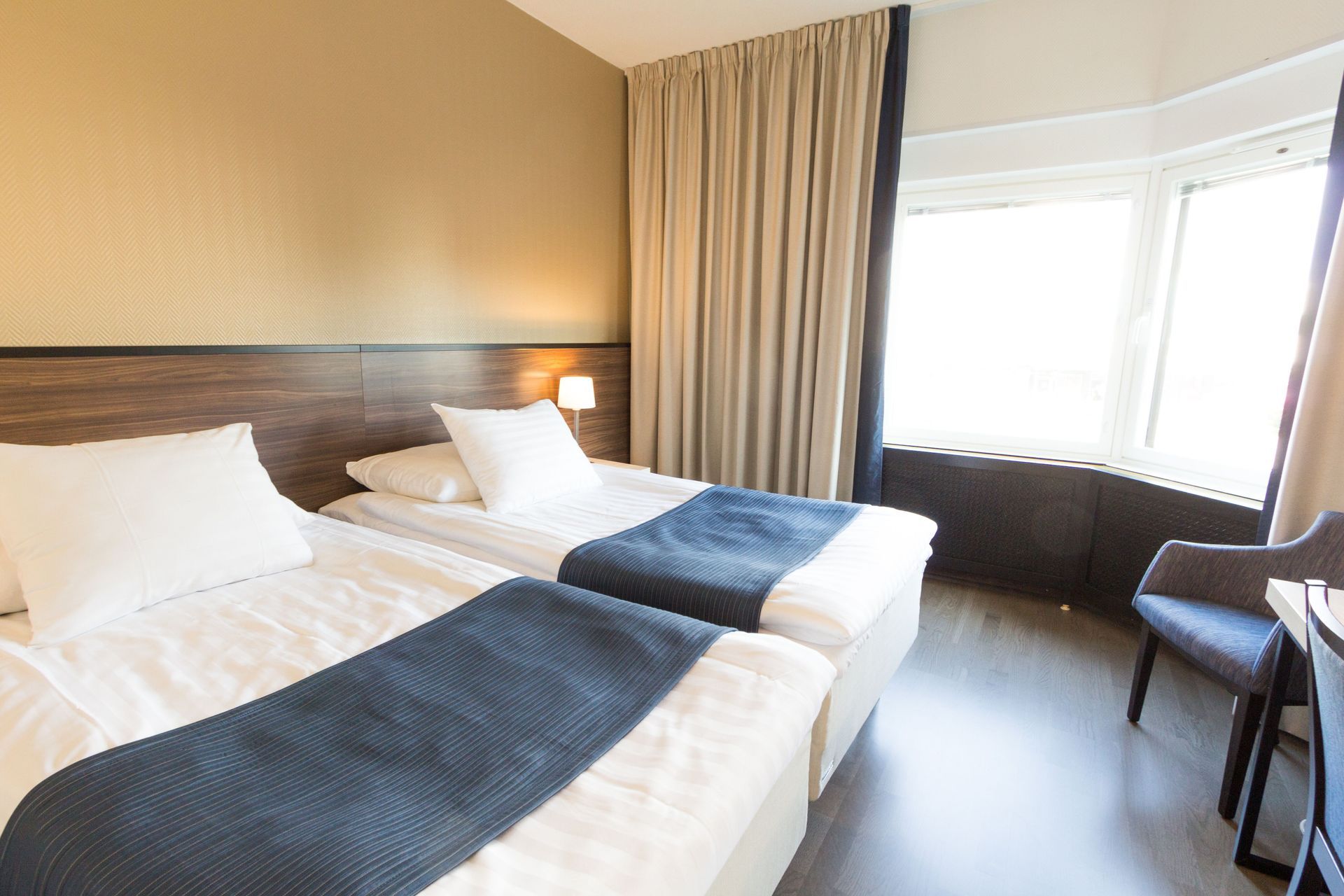 Hotellihuone, jossa on kaksi erillistä sänkyä valkoisilla lakanoilla, tummansinisillä juoksijaliinoilla ja vaaleilla verhoilla peitetty suuri ikkuna.