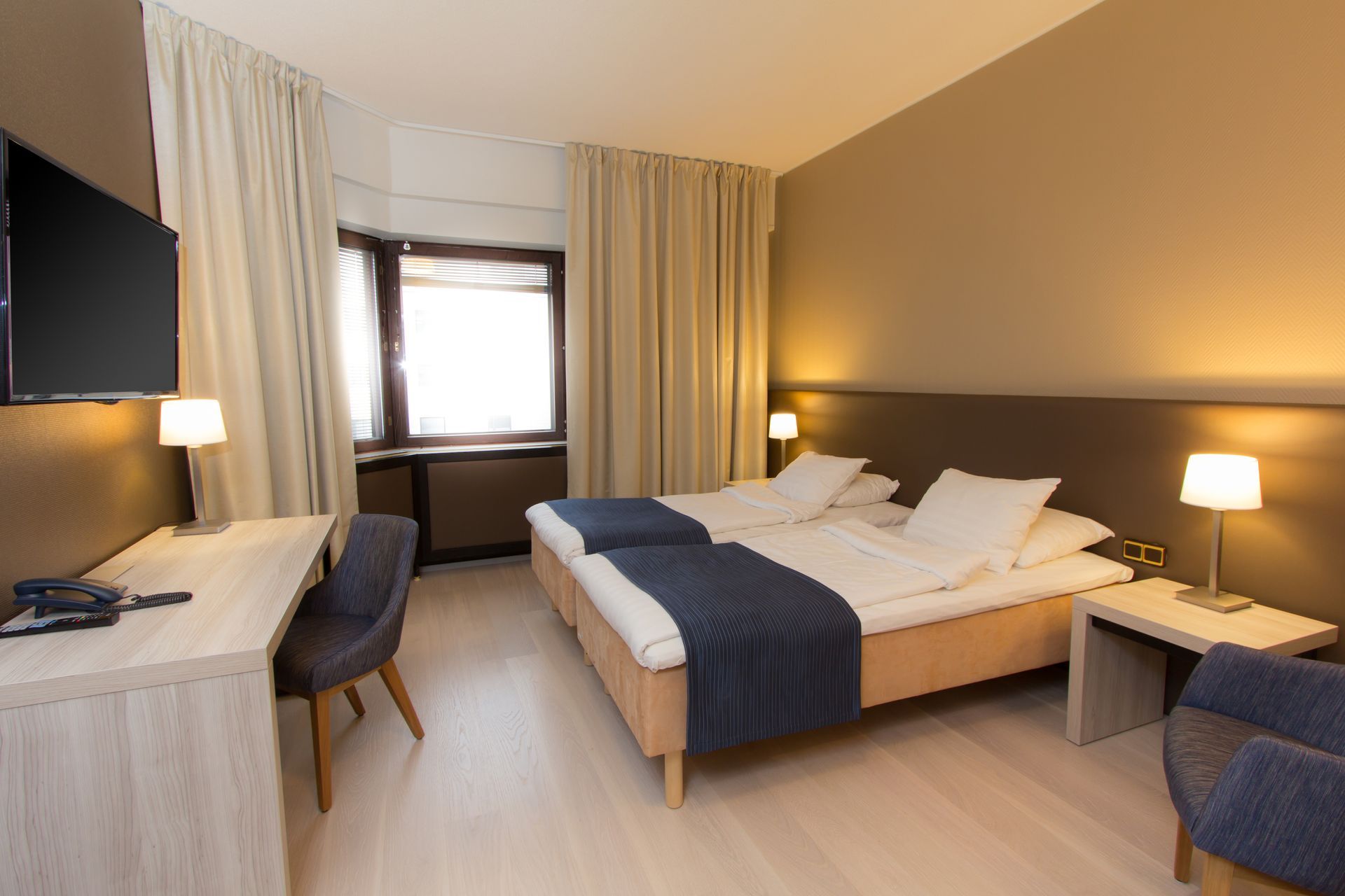 Moderni hotellihuone, jossa on kaksi erillistä sänkyä, työpöytä, televisio ja verhoilla varustettu ikkuna.