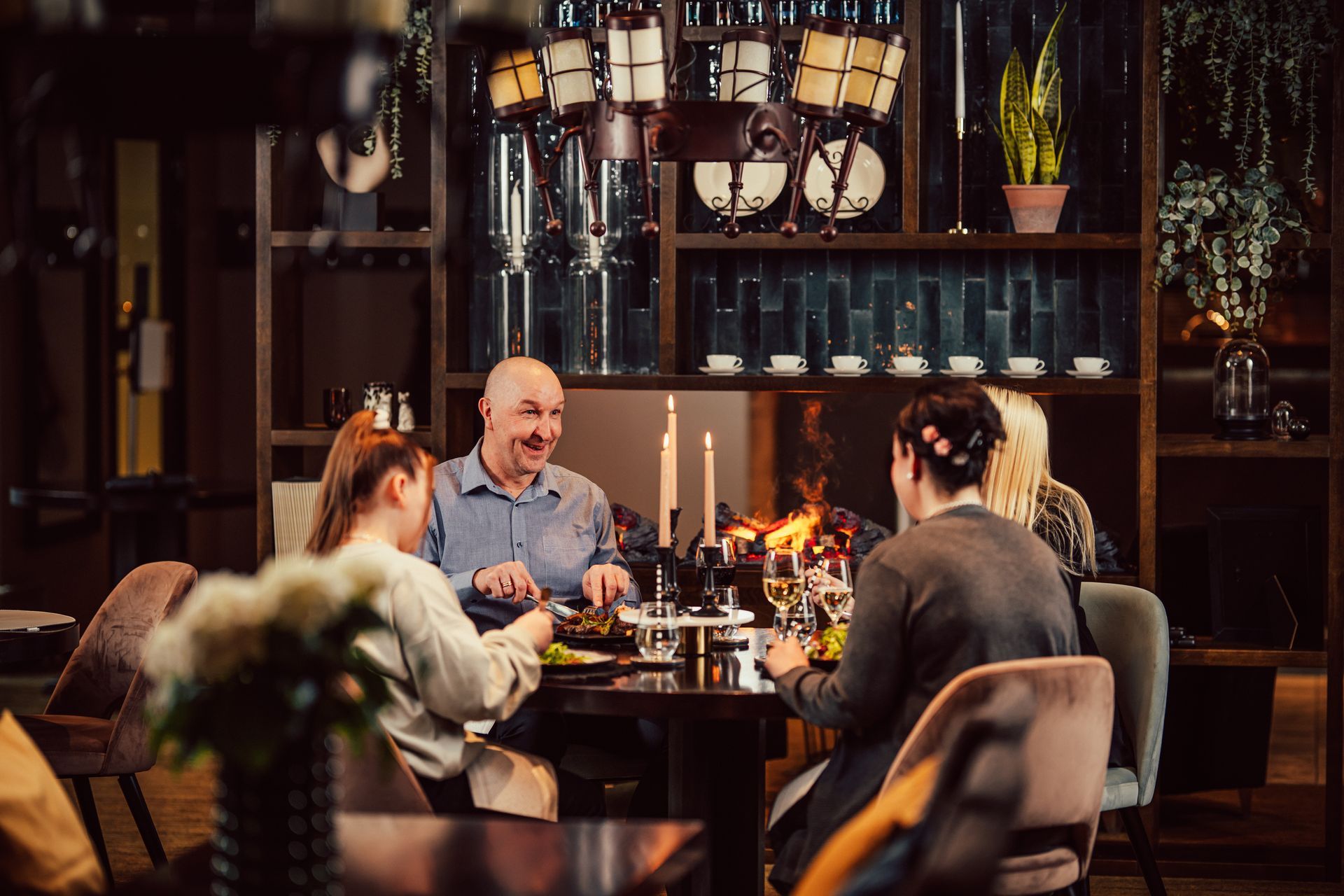 Three people eating at a table in a dimly lit, warm-toned restaurant with a fireplace in the background.