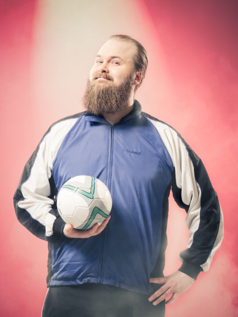 Hattu ja liivi yllään oleva henkilö istuu hymyillen vintage-pinkillä sohvalla sinivihreää, teksturoitua seinää vasten.