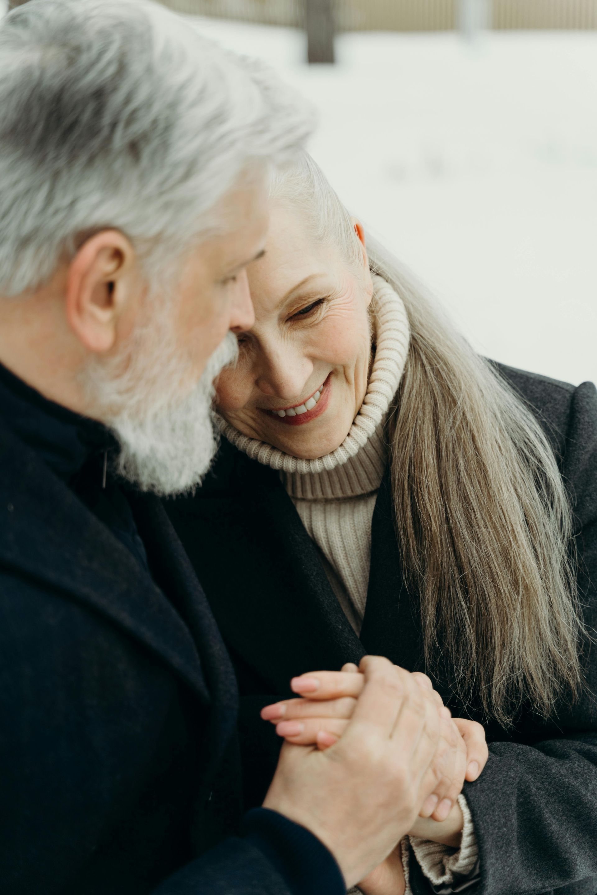Two people with gray hair are smiling warmly while holding hands, wearing coats and a turtleneck sweater in a snowy area.