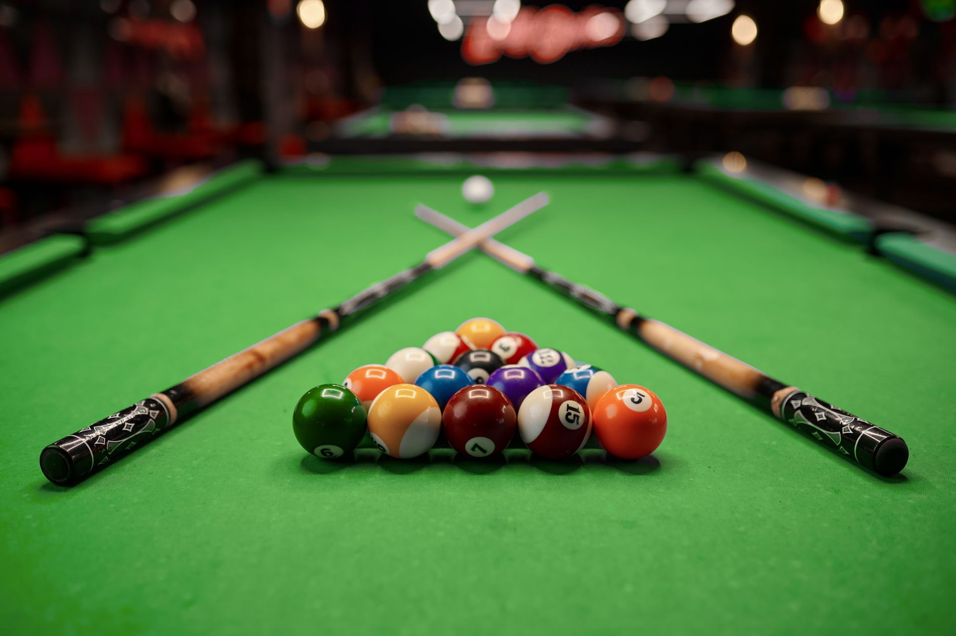 Pool table with balls racked, cues crossed, and white cue ball. Green felt, wooden cues.