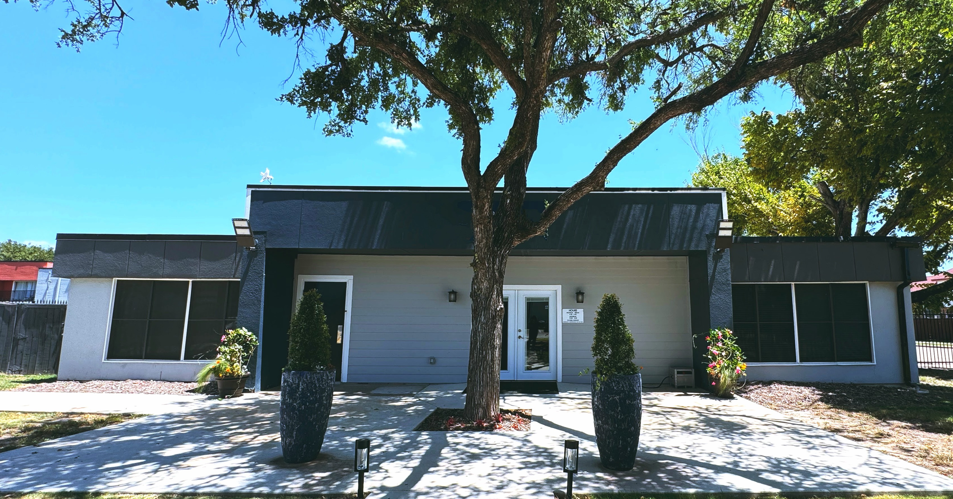 A modern, gray building with a tree in front. Two dark potted plants frame the entrance.