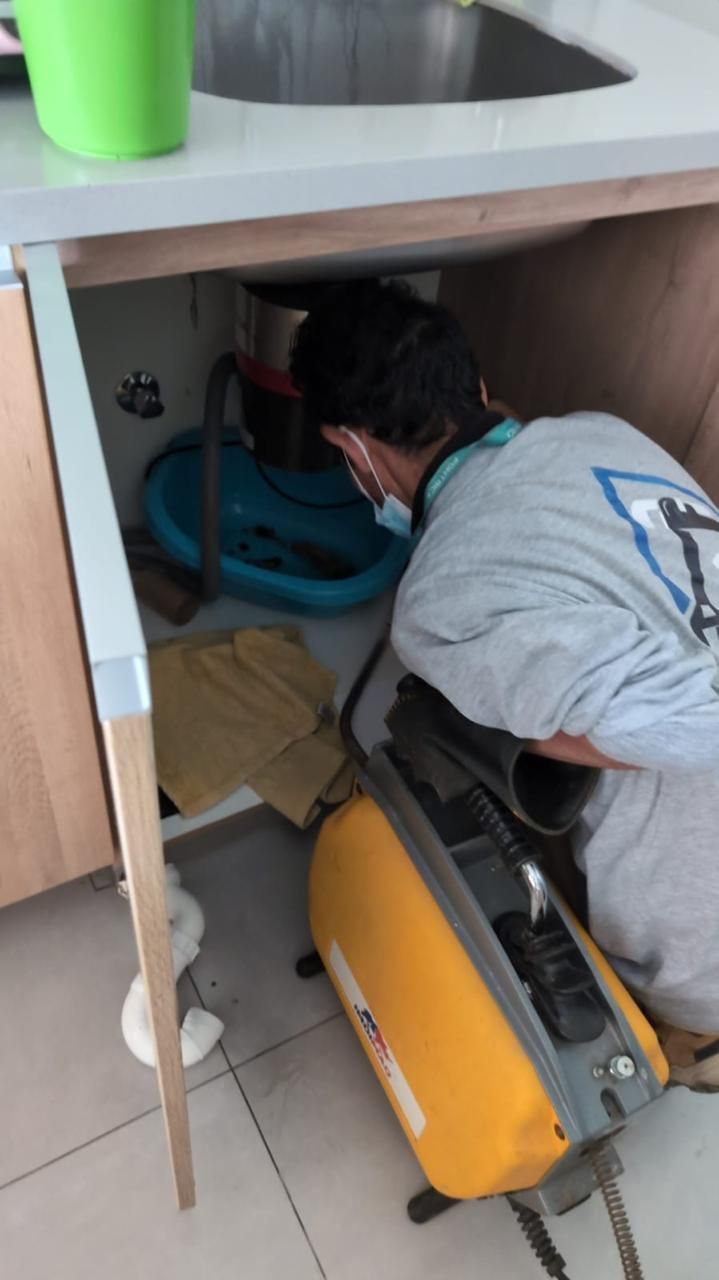 Una persona está agachada debajo de un mueble de fregadero, trabajando en la fontanería con un cubo azul