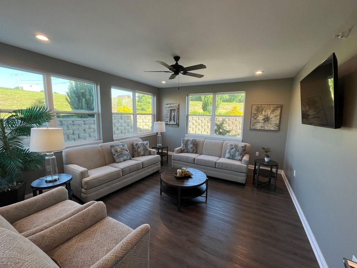 Bright living room with beige sofas, dark wood floor, large windows, ceiling fan, and wall-mounted TV.