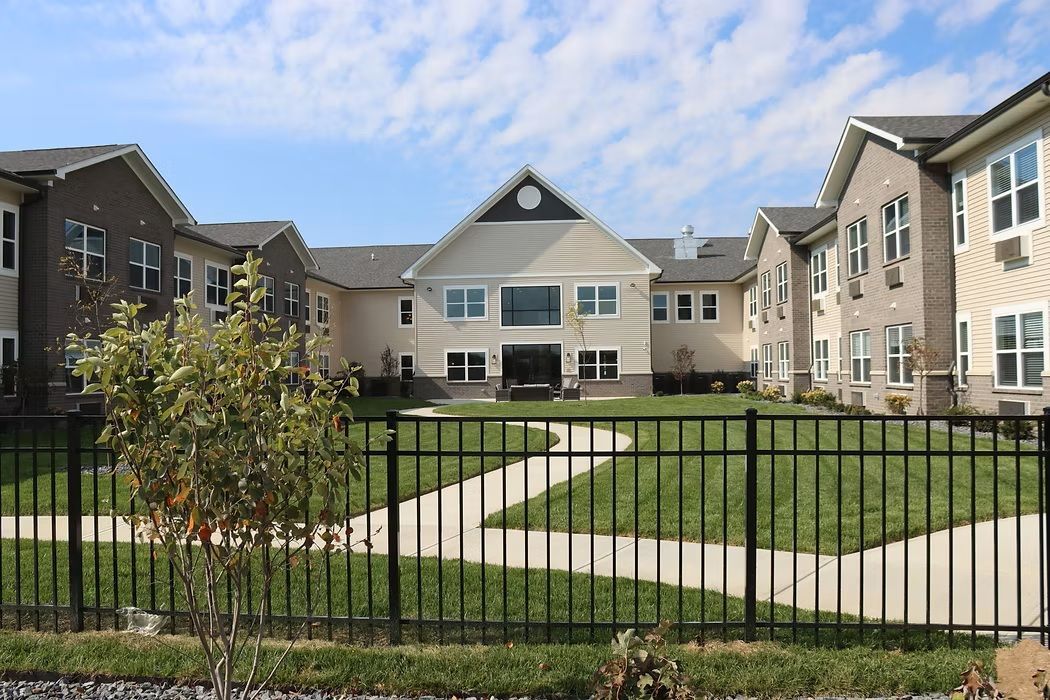 Apartment courtyard with green lawn, black fence, and beige buildings under a partly cloudy sky