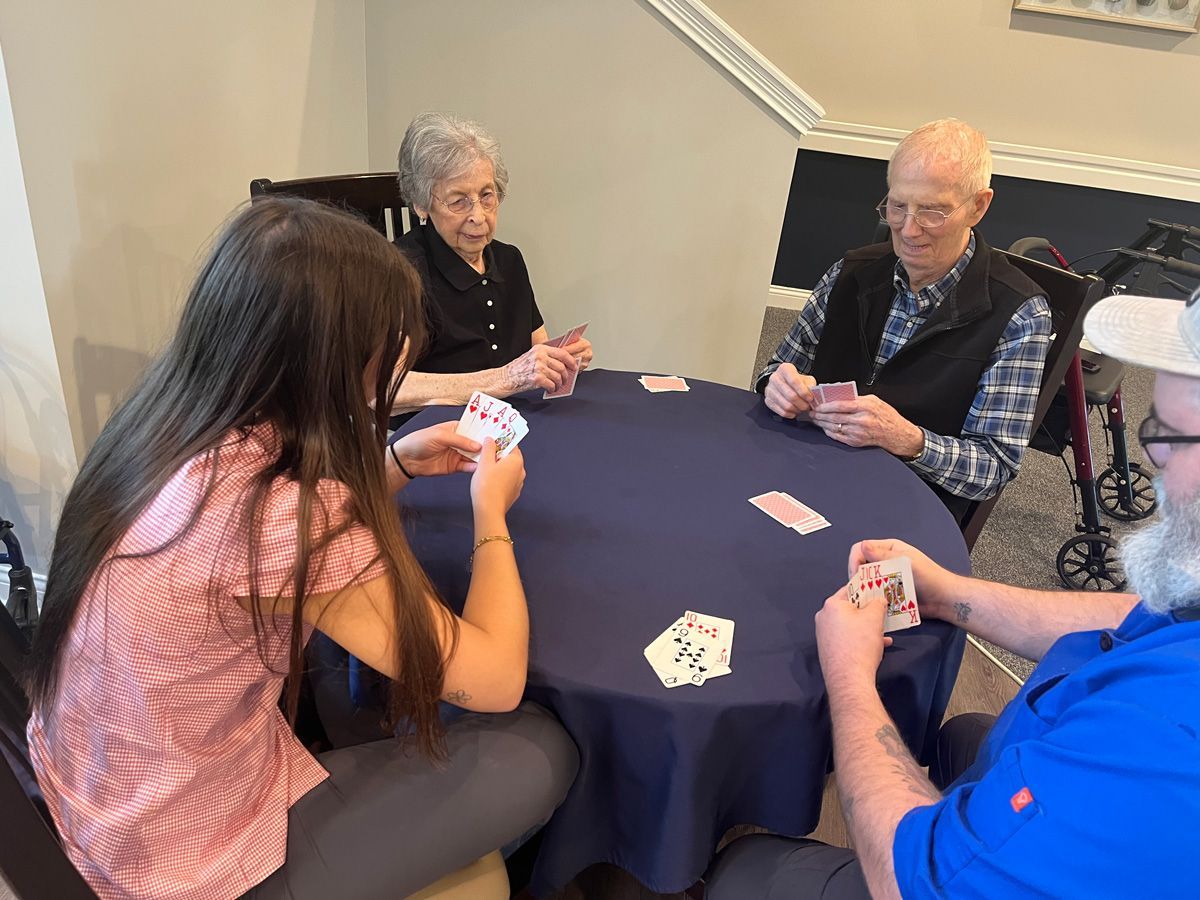 Four people play cards around a navy table in a bright room.