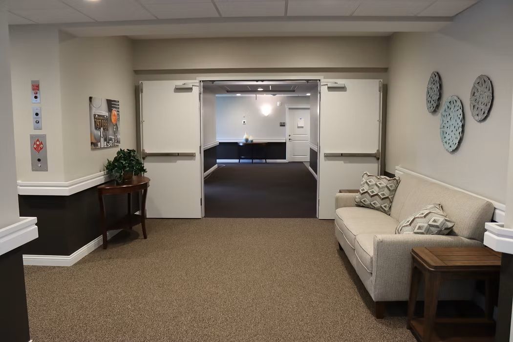 Waiting area with beige chair, side table, wall art, and open double doors to a hallway.