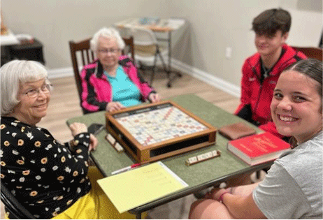Four people playing Scrabble around a table in a bright room, smiling at the camera