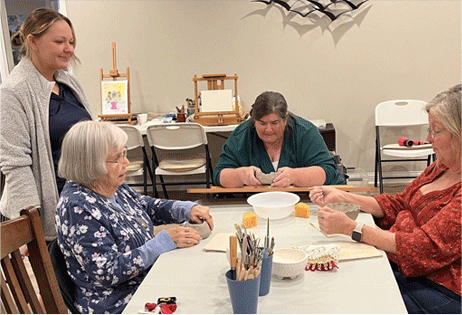 Four people sit around a craft table, drawing with pencils in a bright community room.