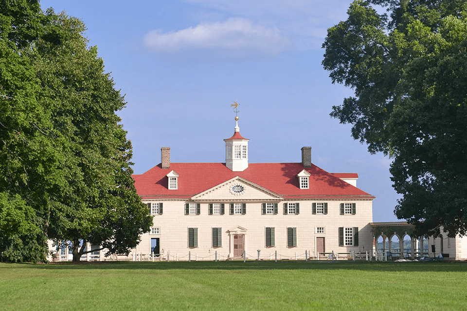 Mount Vernon estate, white mansion with red roof, on a green lawn, framed by trees under blue sky.