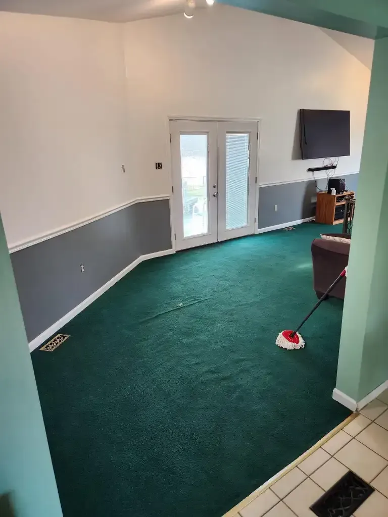 Green carpeted room with white and gray walls, French doors, and a mounted TV. A mop rests on the floor. Green carpeted room with white and gray walls, French doors, and a mounted TV. A mop rests on the floor.