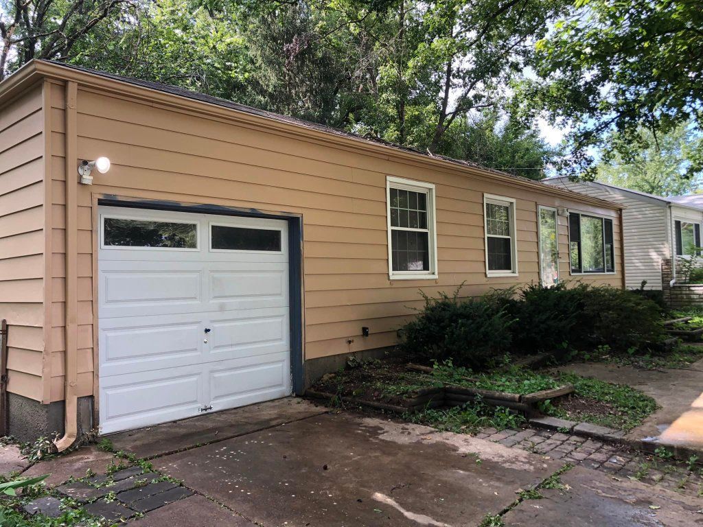 Tan garage with white door and three windows. Bush in front of building.