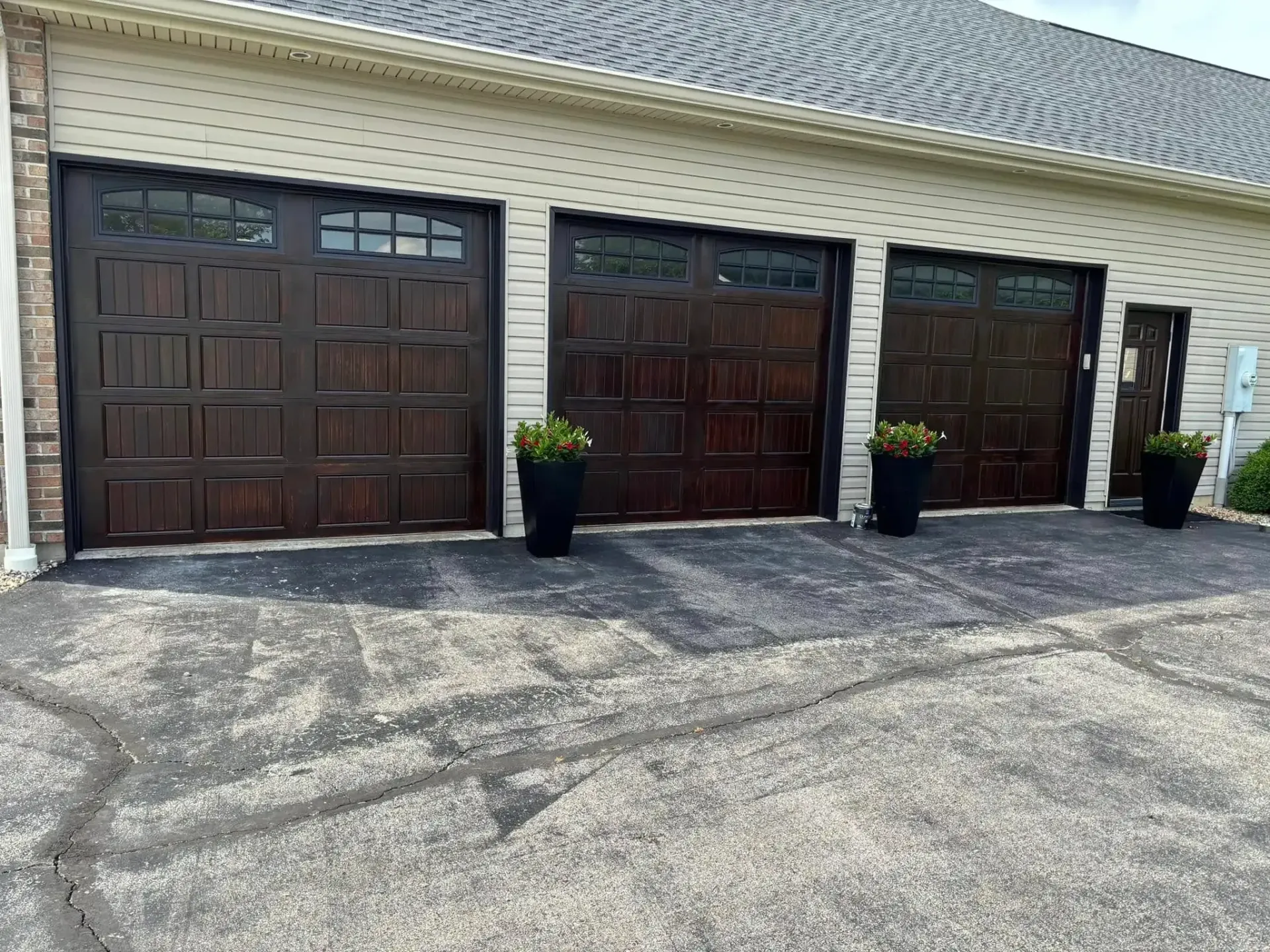 Three dark brown garage doors with decorative windows, potted plants, and an asphalt driveway.